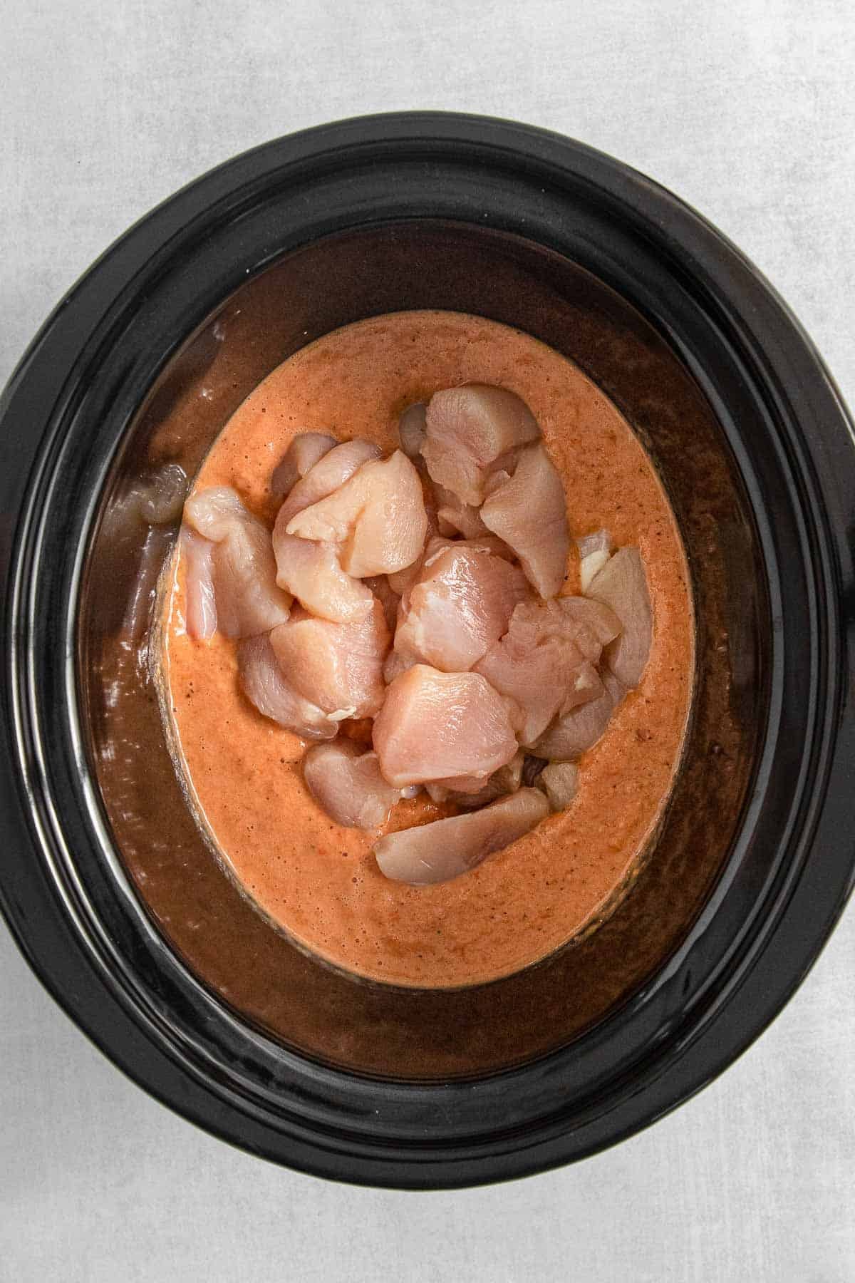 A black crock pot filled with raw chicken in a red cream sauce.