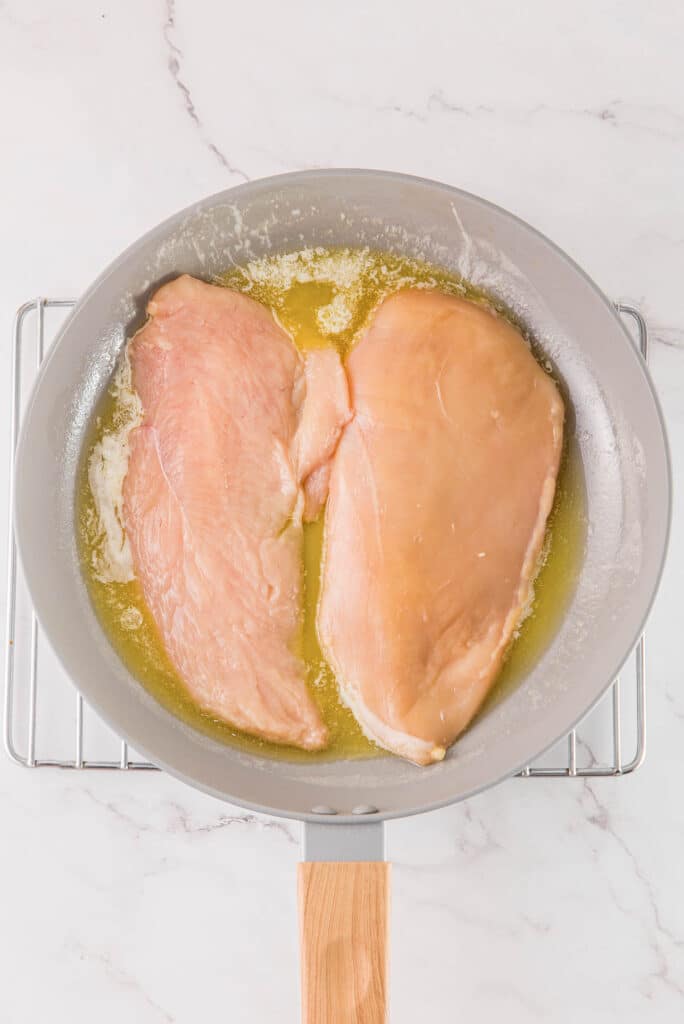 Two raw chicken breasts in a frying pan with melted butter.