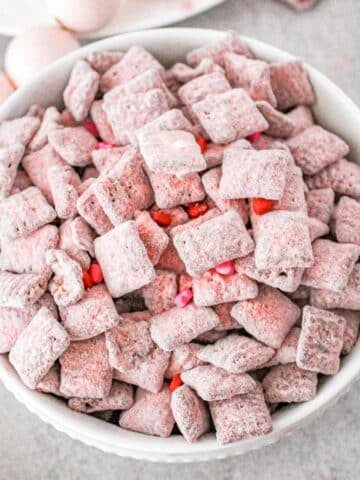white bowl full of strawberry muddy buddy recipe with red candy hearts in it.