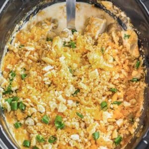 A black slow cooker filled with cheesy chicken and potatoes topped with crushed Ritz crackers being scooped with a big silver spoon.