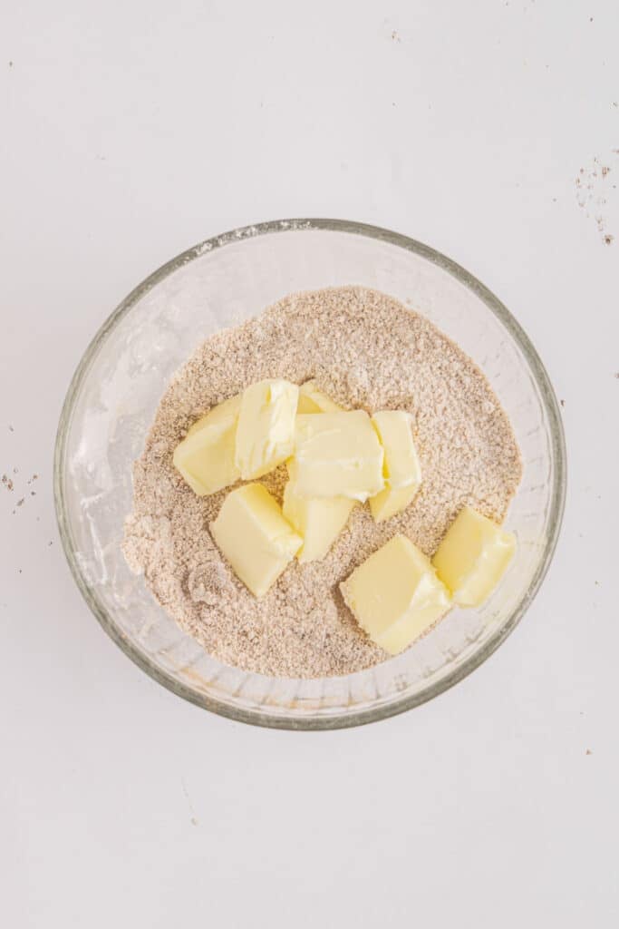 Glass mixing bowl with a mixture of flour, brown sugar, sugar, cinnamon and salt with added butter slices.