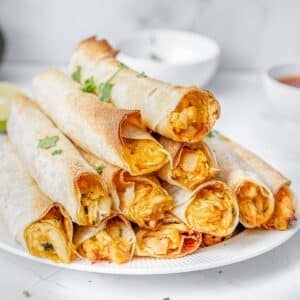 A plate of crispy baked chicken taquitos filled with a chunky, golden filling, garnished with fresh herbs, served with dipping sauces on a white marble surface.