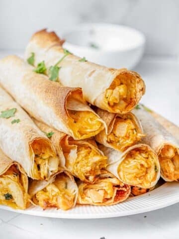 A plate of crispy baked chicken taquitos filled with a chunky, golden filling, garnished with fresh herbs, served with dipping sauces on a white marble surface.