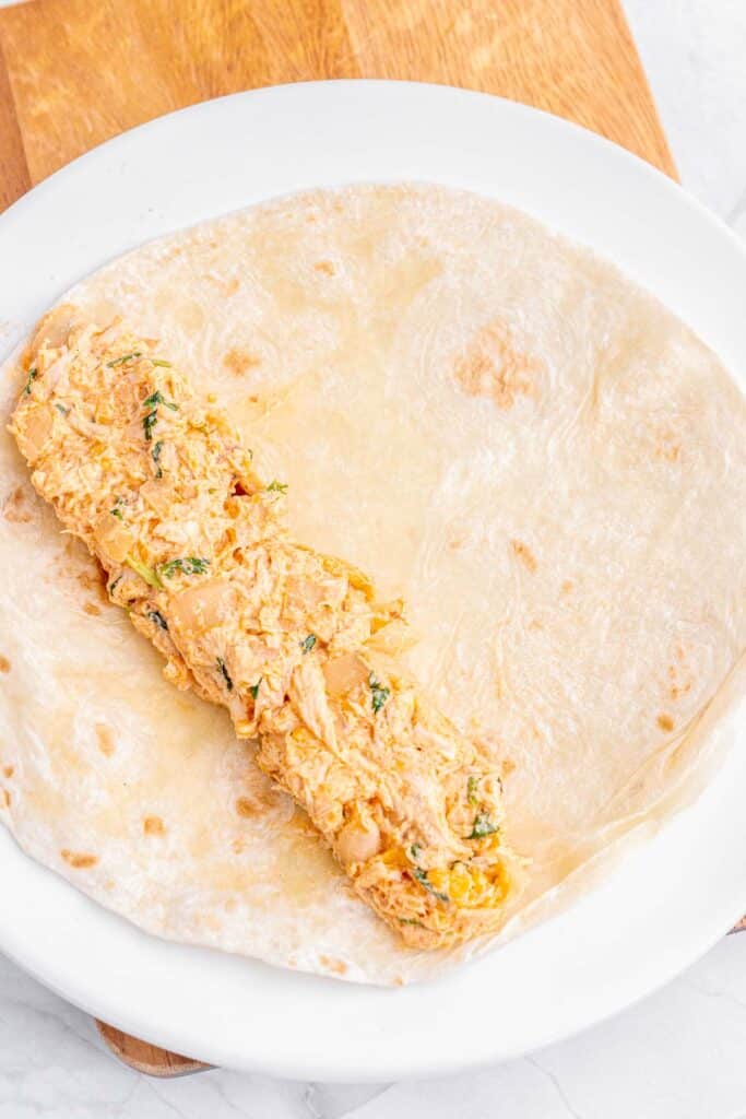 A plate with a large flour tortilla with a serving of creamy, shredded chicken filling, seasoned with herbs, on a wooden board.