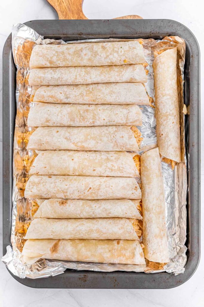 A tray of freshly rolled chicken taquitos laying neatly on a foil lined baking sheet and ready to be cooked.