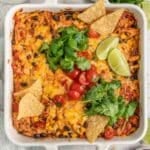 A colorful mexican casserole in a white dish, topped with melted cheese, tomatoes, cilantro, and lime slices, served with tortilla chips on the side.