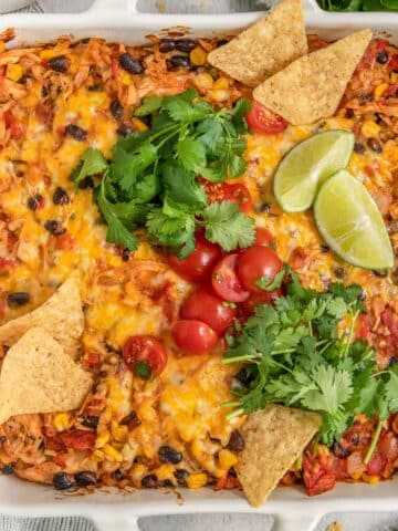 A colorful mexican casserole in a white dish, topped with melted cheese, tomatoes, cilantro, and lime slices, served with tortilla chips on the side.