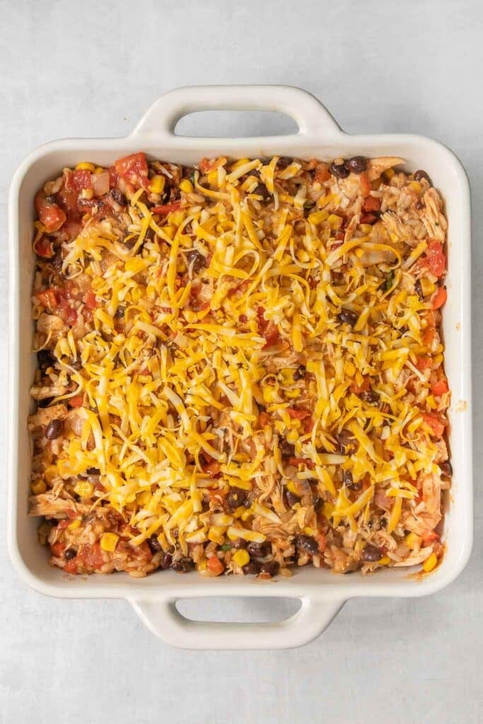 A square baking dish filled with a colorful baked casserole topped with melted cheddar cheese, featuring layers of beans, rice, tomatoes, and peppers.