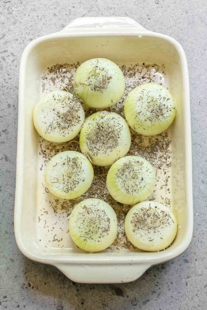 eight peeled onions with tops sliced off, placed in a baking dish, sprinkled with dried herbs, ready to be roasted.