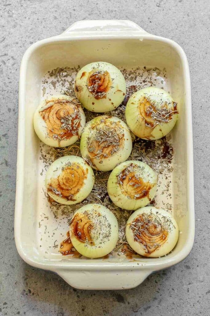 Eight peeled onions seasoned with spices and a drizzle of balsamic vinegar, neatly arranged in a white rectangular baking dish on a grey surface.