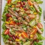 A colorful Mexican grilled chicken salad with sliced tomatoes, cucumbers, corn, and kidney beans on a bed of lettuce, garnished with lime wedges.