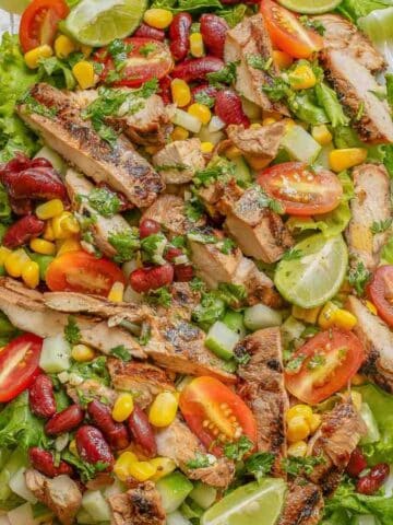 A colorful Mexican grilled chicken salad with sliced tomatoes, cucumbers, corn, and kidney beans on a bed of lettuce, garnished with lime wedges.