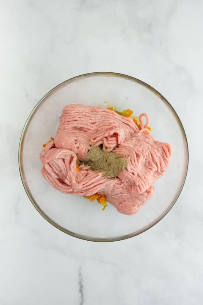 Raw ground turkey with spices and herbs in a glass bowl on a marble countertop, ready for mixing.
