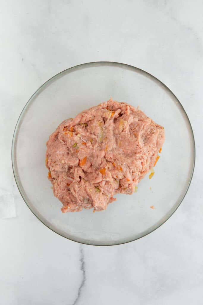 A bowl of raw ground turkey mixed with diced vegetables on a marble surface.
