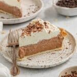 A slice of chocolate cream pie topped with whipped cream and chocolate shavings, served on a white plate with a fork beside it.