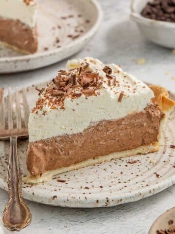 A slice of chocolate cream pie topped with whipped cream and chocolate shavings, served on a white plate with a fork beside it.