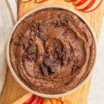 A bowl of chocolate hummus topped with chocolate chunks surrounded by sliced apples.``
