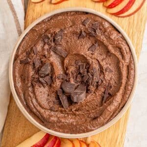 A bowl of chocolate hummus topped with chocolate chunks surrounded by sliced apples.``