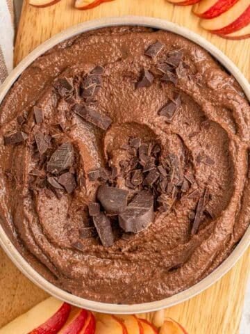 A bowl of chocolate hummus topped with chocolate chunks surrounded by sliced apples.``