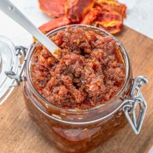 A close-up of a glass jar filled tomato jam placed on a wooden board with a spoon inserted.