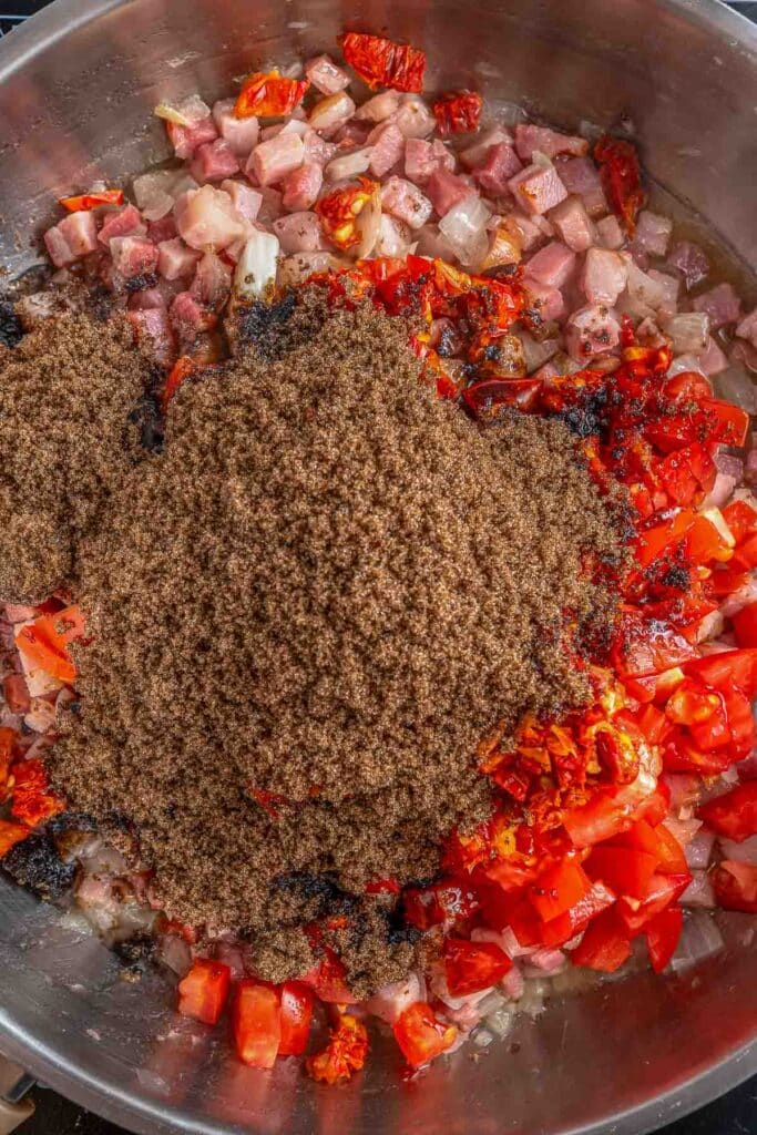 A large pan filled with a mixture of chopped onions, diced bacon, and chopped tomatoes with brown sugar sprinkled on top.