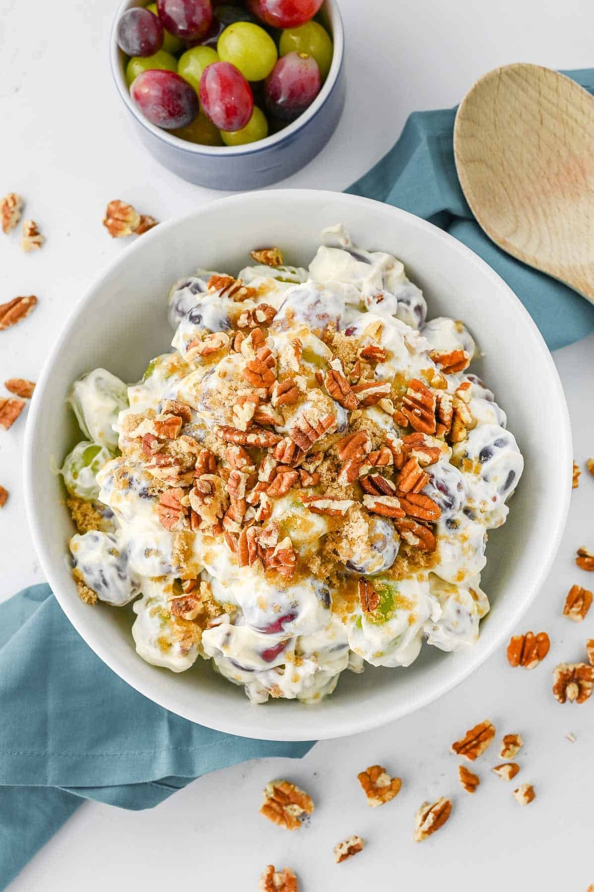 A bowl of creamy grape salad garnished with chopped pecans is placed on a teal cloth. Scattered pecans surround the bowl. A wooden spoon lies nearby.