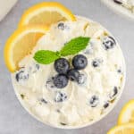 A white bowl of creamy lemon fluff topped with fresh blueberries and a sprig of mint, surrounded by lemon slices.