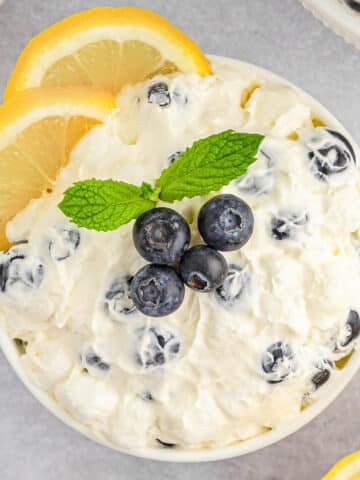 A white bowl of creamy lemon fluff topped with fresh blueberries and a sprig of mint, surrounded by lemon slices.