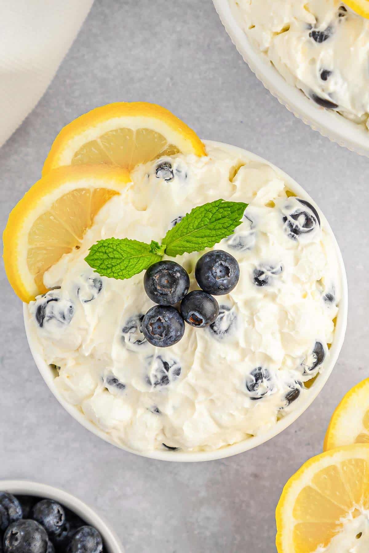 A white bowl of creamy lemon fluff topped with fresh blueberries and a sprig of mint, surrounded by lemon slices.