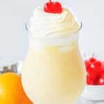 A stemmed glass of orange milkshake topped with whipped cream and a cherry, with an orange and a bowl of cherries in the background. Text at the top reads, "Easy Orange Milkshake.