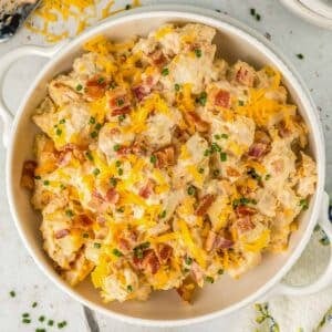 A white bowl of ranch potato salad topped with shredded cheese, crispy bacon bits, and chopped chives.