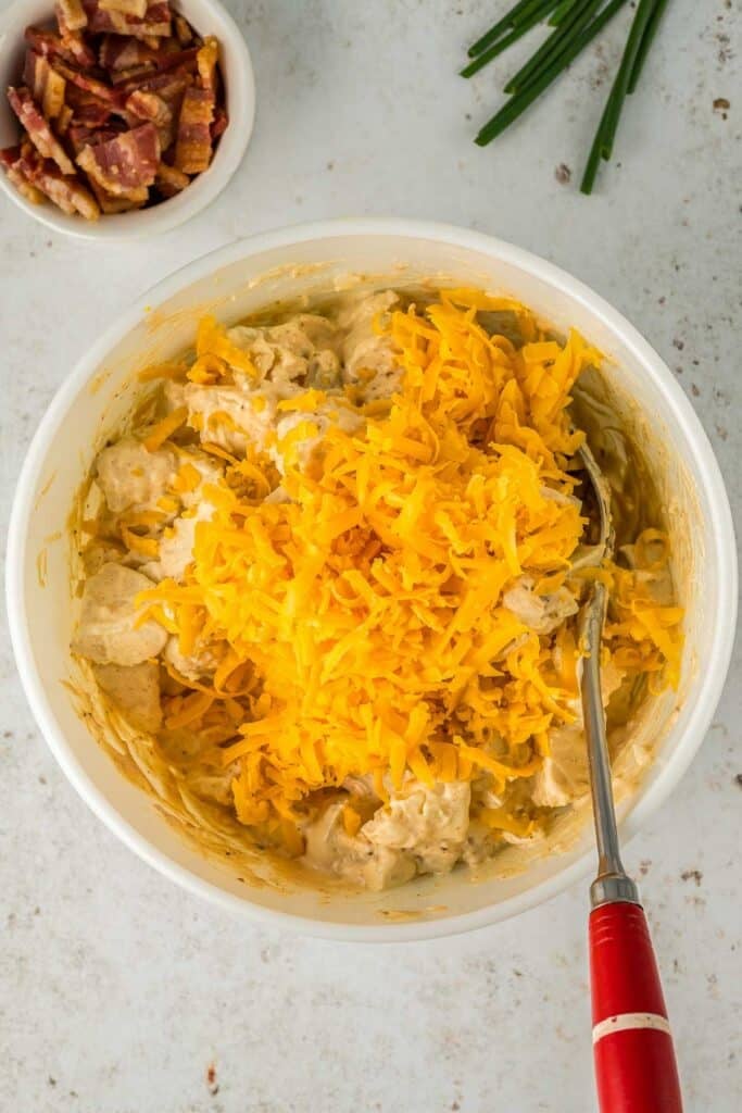 A bowl of potatoes covered in a ranch dressing mixture topped with shredded cheddar cheese, with a spoon with a red handle is in the bowl.