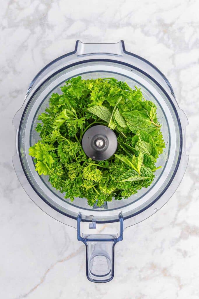 Top view of fresh green herbs, including parsley and mint, in a food processor on a white marble countertop.