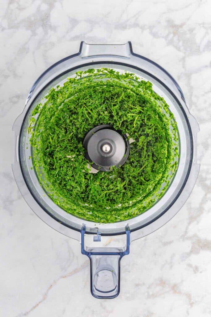 Top-down view of finely chopped parsley and mint in a food processor bowl.