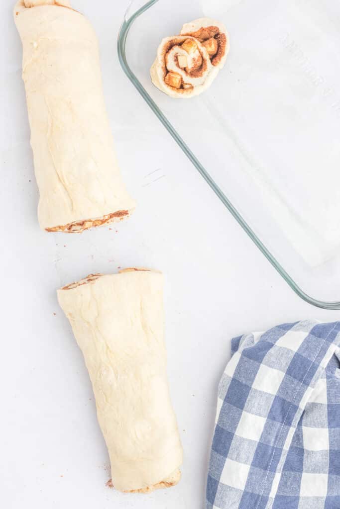 Two unbaked apple cinnamon roll logs and one sliced roll next to a glass baking dish and a blue-and-white checkered cloth.