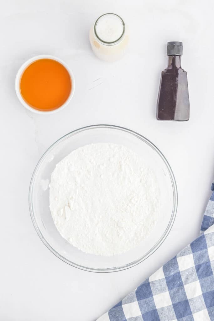 A glass bowl of flour, small bowl of maple syrup, bottle of vanilla extract, and cup of milk.