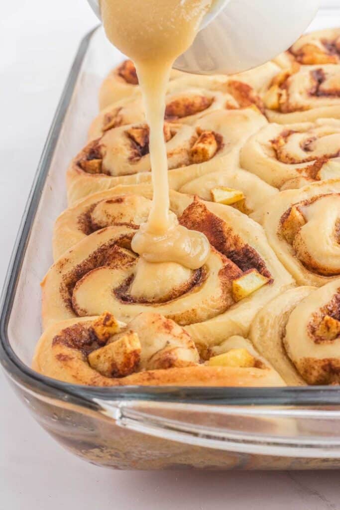 Maple glaze is being poured over freshly baked apple cinnamon rolls in a rectangular glass baking dish.