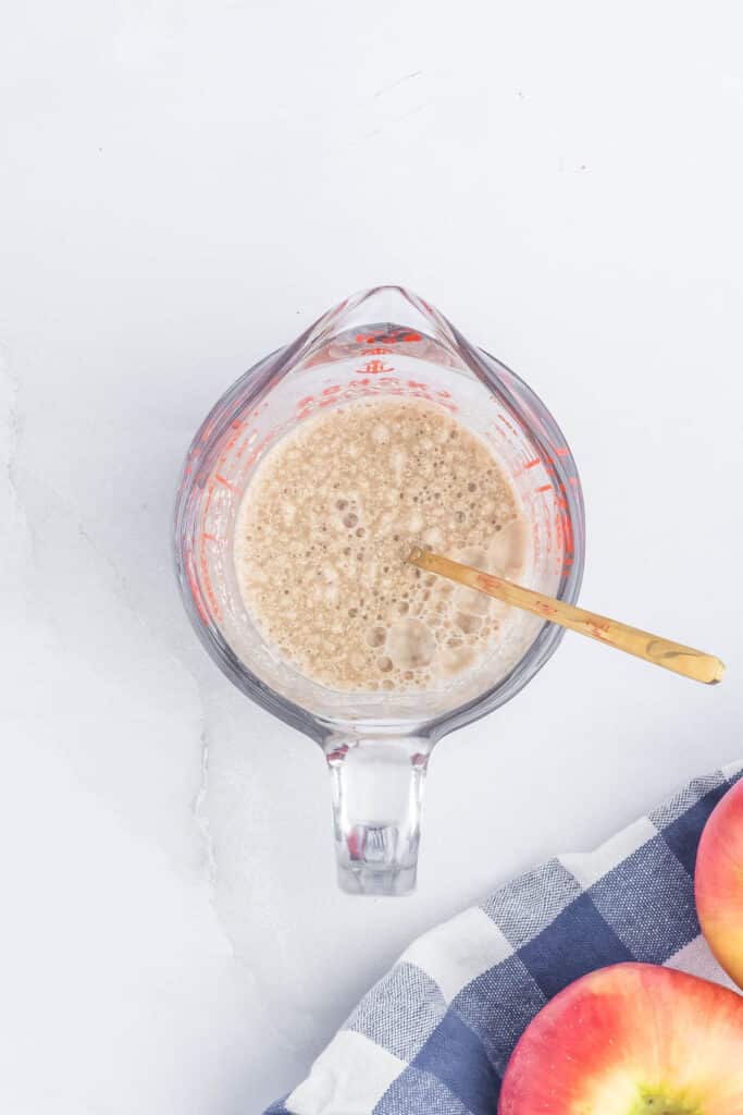 Brown sugar, yeast and water mixed together in a measuring cup next to 2 apples on a white and blue checkered towel.