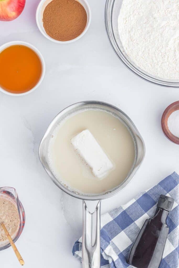 A saucepan containing milk and butter on a stovetop next to 2 apples on a white and blue checkered towel.