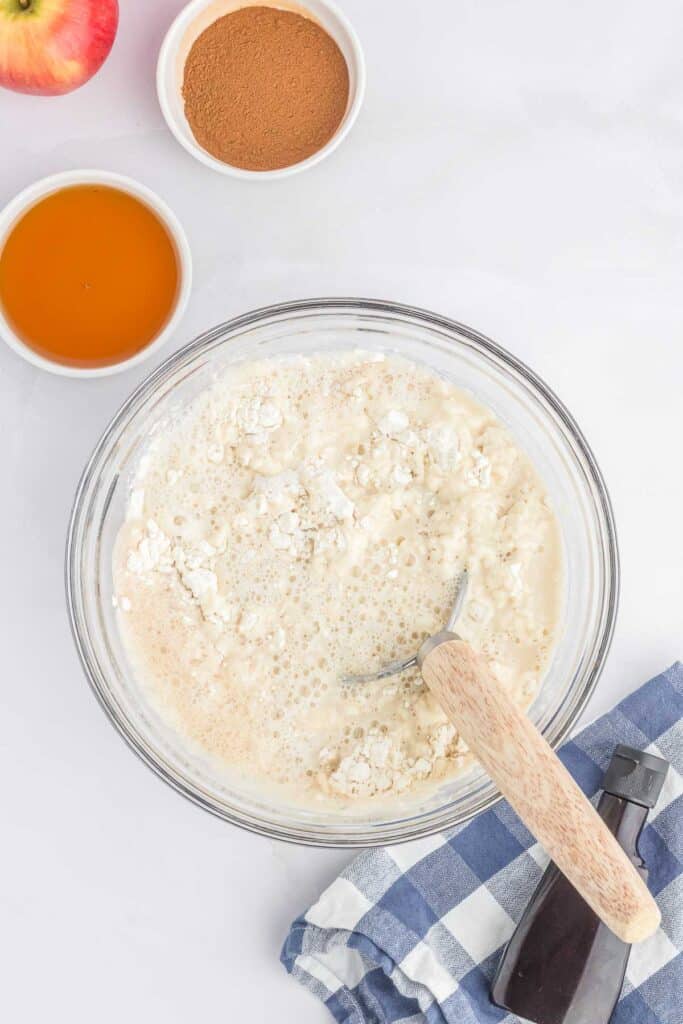 A clear mixing bowl containing the flour mixture with the butter and sugar mixture and the yeast and sugar mixture poured over top.