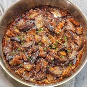A skillet filled with cooked chinese eggplant slices in a savory sauce, garnished with sesame seeds and chopped green onions.