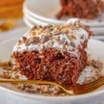 Close-up of a chocolate caramel cake slice on a white plate, topped with cool whip and crushed heath bar bits, with a fork on the side.