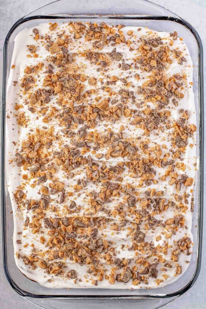 Chocolate caramel cake with cool whip spread evenly overtop in a large clear glass baking dish with heath bar crumbled covering.