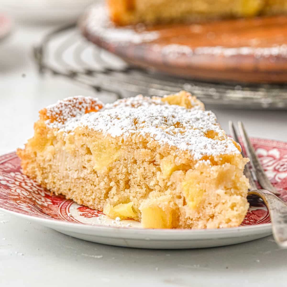 A slice of french apple cake dusted with powdered sugar on a red and white patterned plate with a fork beside it.