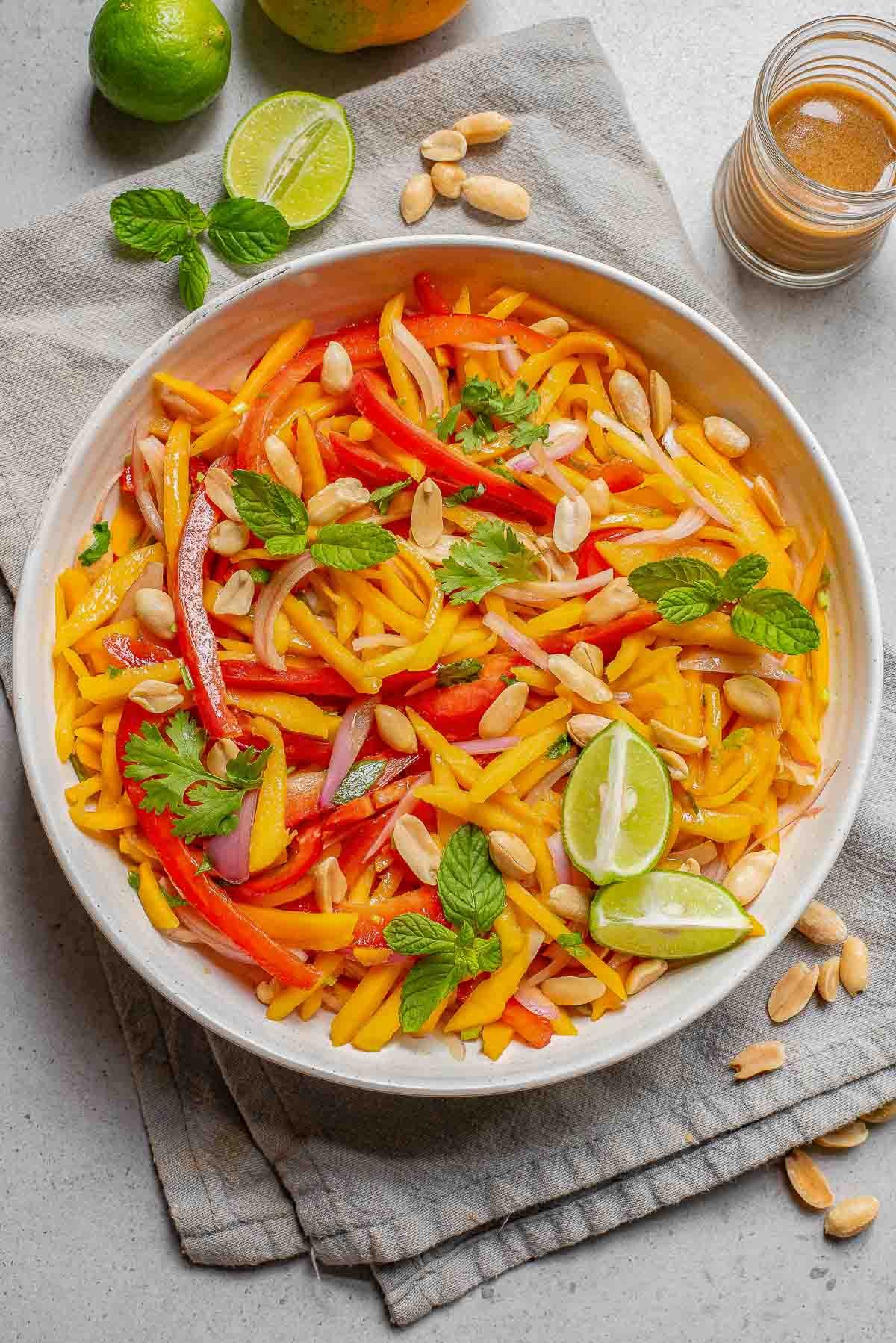 Mango salad with sliced red peppers, fresh mint, lime wedges, and roasted peanuts in a large white bowl.