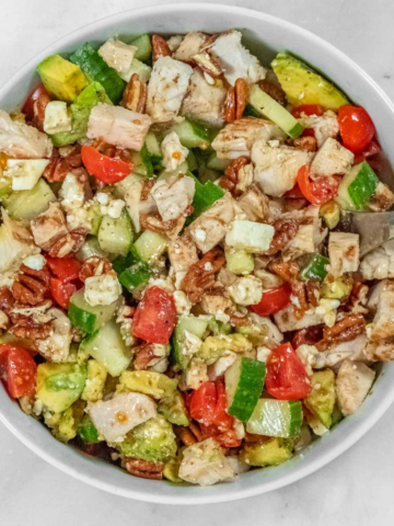 A bowl filled with a salad consisting of diced chicken, cucumbers, tomatoes, avocado, almonds, and cheese crumbles.