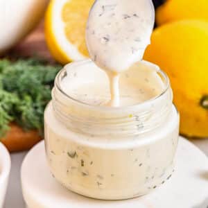 A glass jar filled with creamy lemon aioli, with a spoon being lifted out, and fresh lemons in the background.