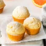 Three orange muffins, dusted with sugar, are placed on parchment paper on top of a tray.