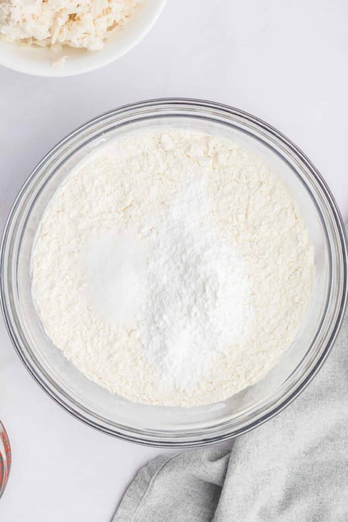A clear glass bowl filled with flour, baking powder and salt on a white surface.