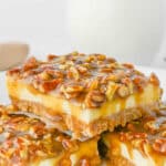 Image of pecan pie cheesecake bars, with a layer of caramel and pecan pieces on top of a creamy cheesecake layer and a graham cracker crust, displayed on a white plate in front of a glass of milk.